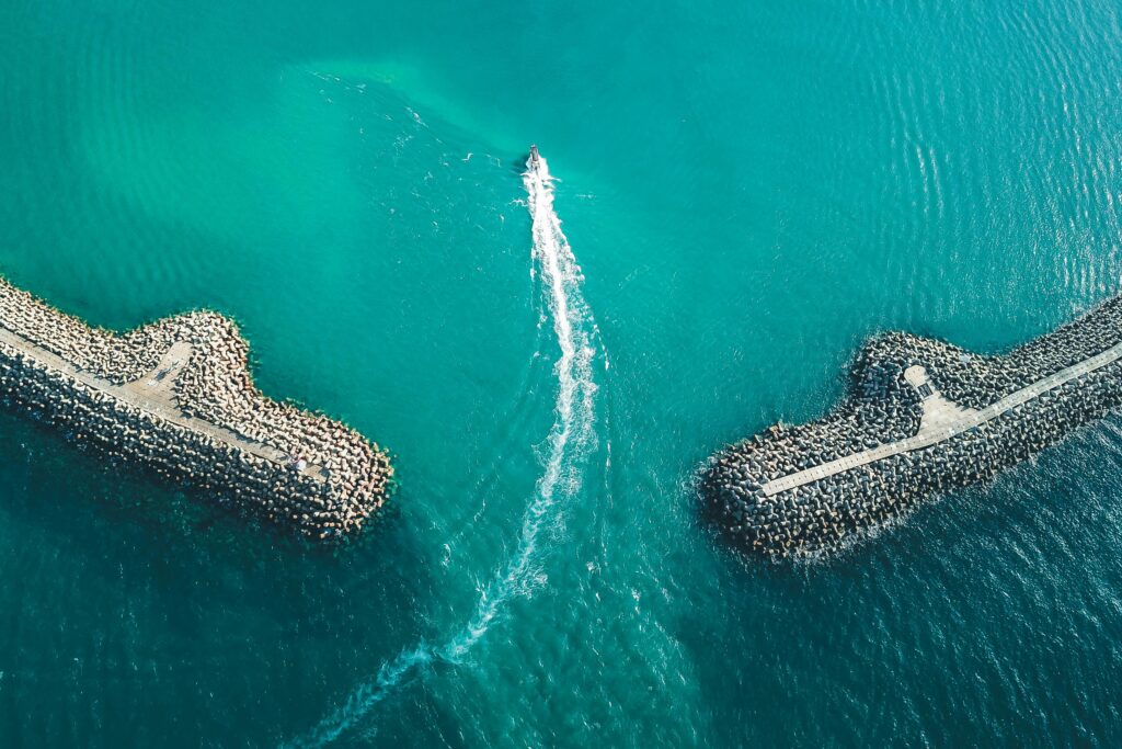 Drone shot capturing a boat traveling between two stone breakwaters in a serene ocean setting.